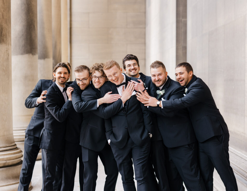 Groomsmen and bridesmaids joyfully hugging the groom in a playful moment at Mellon Institute Columns.