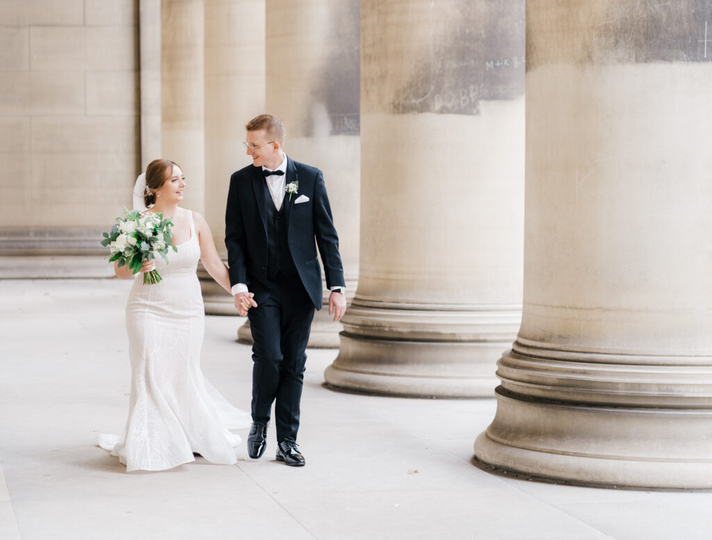 Bride and groom holding hands while walking together in front of Mellon Institute Columns on their wedding day."