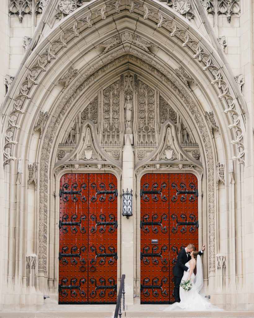 Bride and groom sharing a romantic kiss outside in front of the iconic red doors of Heinz Chapel.